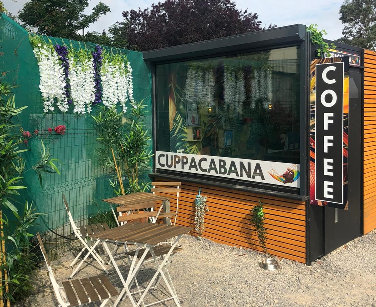 The Cuppacabana kiosk beside Donabate train station, with its orange timber cladding, the CUPPACABANA sign and vertical COFFEE board, and two outdoor bistro tables