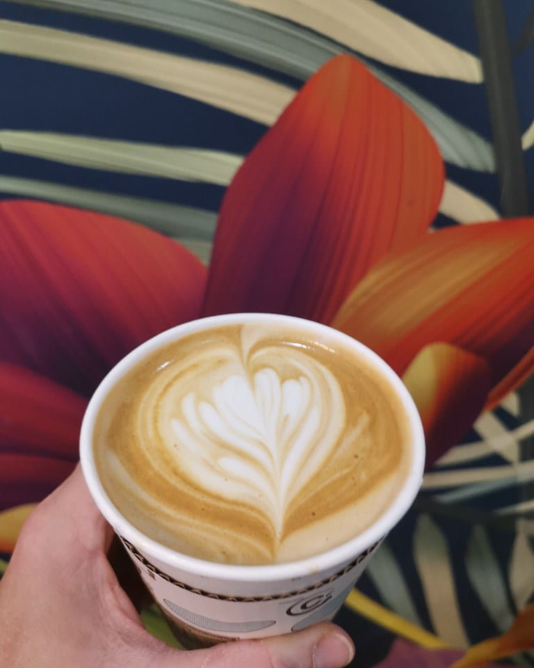 A takeaway cup of latte art held up against the tropical mural inside the kiosk